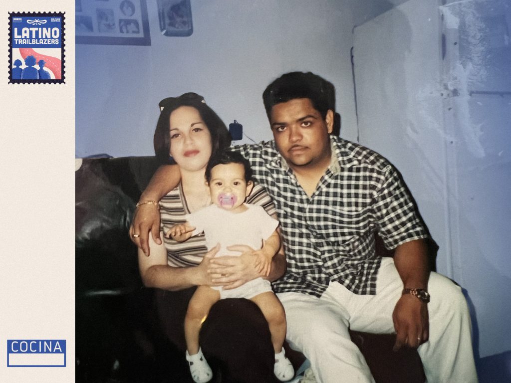 Henna Sharee, part of Latino Trailblazers, as a baby posing with her parents sitting on a couch. Henna is sitting at her mother's lap.