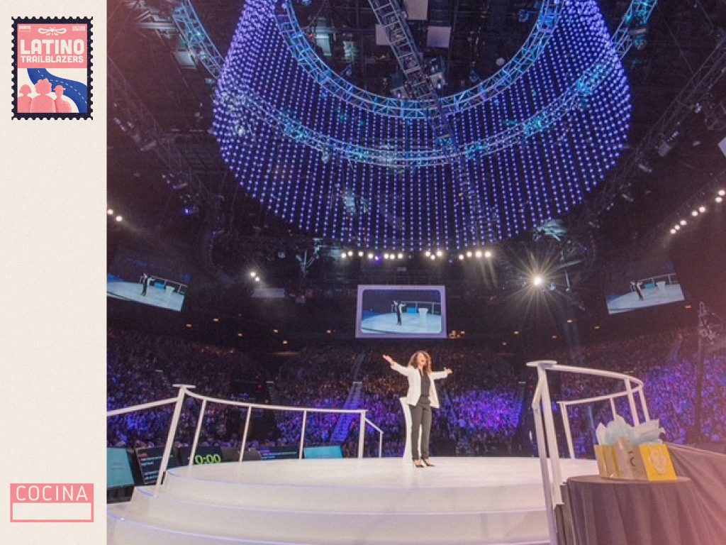 Claire Risoli, part of Latino Trailblazers, in a conference. She's standing in a central, round stage inside a closed stadium, sorrounded by stands filled with people. Photo courtesy of Claire Risoli.