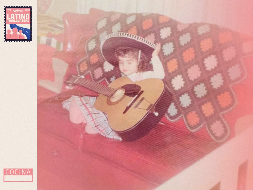 Claire Risoli, part of Latino Trailblazers, as a small child. She's sitting in a couch, wearing a traditional hat and holding a guitar that looks just as big as her. Photo courtesy of Claire Risoli.
